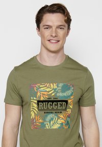 Alyvuogių spalvos medvilninis marškinėliai trumpomis rankovėmis, papuošti spalvinga tropine lapo grafika ir užrašu „RUGGED KOROSHI DENIM“.