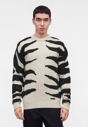 Jeune homme portant un pull beige avec des motifs audacieux de rayures noires de tigre, debout devant un fond blanc uni.