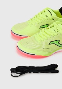 Neon gula sportskor med en texturerad yta, svarta och rosa accenter, samt en platt sula. Svarta skosnören ligger bredvid skorna.