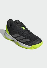 Baskets de sport noires avec une tige en mesh, accent vert, logo à trois bandes grises et semelle blanche. Détails texturés sur les orteils et la semelle.