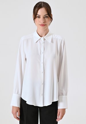 Camicia bianca a maniche lunghe realizzata in tessuto morbido, con colletto classico, chiusura con bottoni e orlo leggermente curvo.