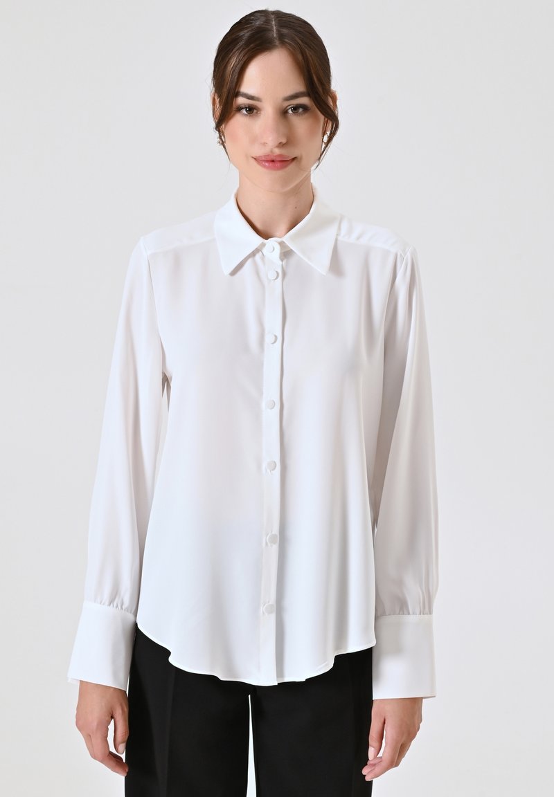 Camicia bianca a maniche lunghe realizzata in tessuto morbido, con colletto classico, chiusura con bottoni e orlo leggermente curvo.
