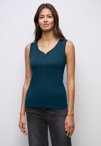 Top senza maniche di colore teal con scollatura a V e due bottoni sul petto, realizzato in un tessuto liscio e elasticizzato; abbinato a jeans grigio scuro.
