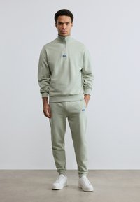 Άνδρας που φοράει ανοιχτοπράσινο φούτερ με φερμουάρ και αντίστοιχο παντελόνι φόρμας με λευκά αθλητικά παπούτσια, στέκεται μπροστά σε απλό ανοιχτό γκρι φόντο.