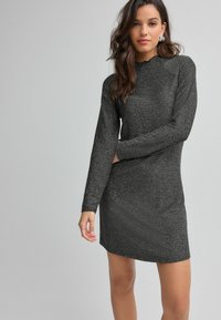Rochie neagră, sclipitoare, tip shift, cu mâneci lungi, gât înalt și o siluetă ajustată. Textură netedă cu o ușoară strălucire.
