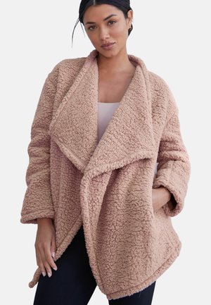 Vrouw draagt een zachte, oversized sherpa-jas in stoffig roze met een brede kraag, één hand in de zak, tegen een effen lichte achtergrond.