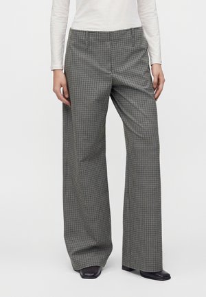 Personne portant un pantalon large à carreaux noir et blanc, des chaussures noires à bout carré et un haut blanc à manches longues, debout devant un fond uni.