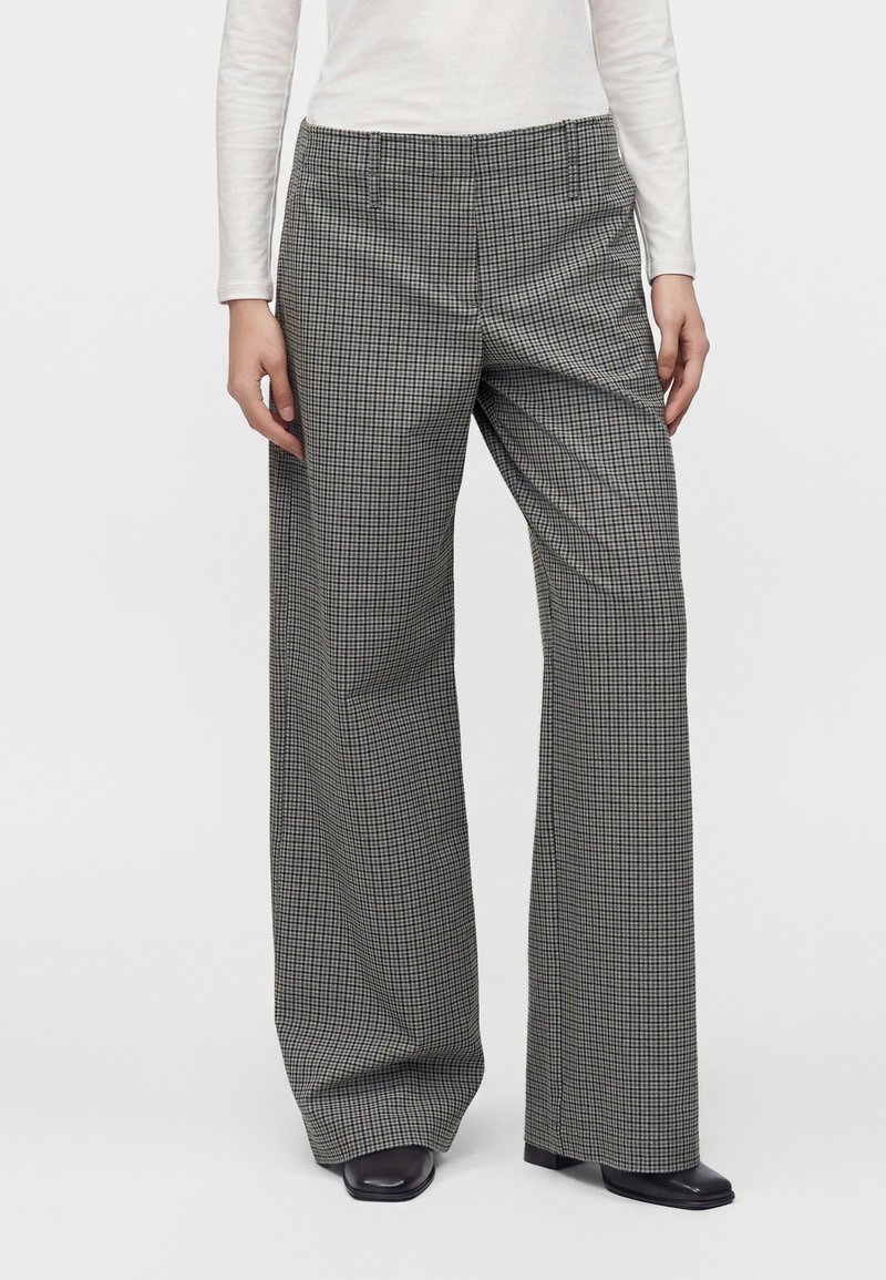 Personne portant un pantalon large à carreaux noir et blanc, des chaussures noires à bout carré et un haut blanc à manches longues, debout devant un fond uni.