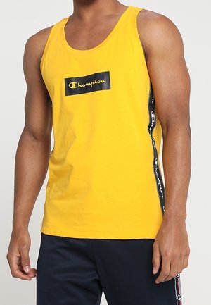 Man draagt een geel Champion mouwloos shirt met logo op de borst en een donkere broek met logostreep aan de zijkant.