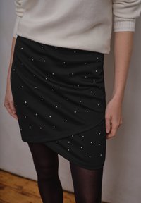 Czarna mini spódniczka z marszczonym wzorem, ozdobiona srebrnymi ćwiekami, noszona z czarnymi rajstopami. Miękka tkanina i teksturowane wykończenie.