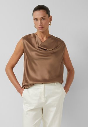 Frau mit zurückgekämmtem Haar, die goldene Creolen trägt, ein braunes ärmelloses Satintop und weiße hoch taillierte Hosen mit Händen in den Taschen.