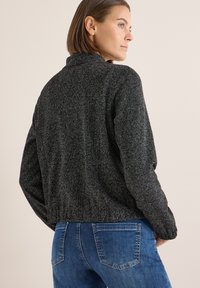 Svart och grå texturerad bomberjacka med ståkrage, elastiska muddar och linning, i kombination med blå jeans.