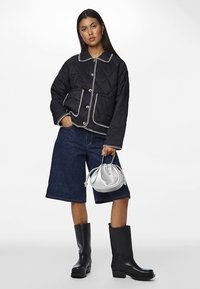 Casaco preto acolchoado com detalhes em branco, botões grandes e bolsos, combinado com calções de ganga escuros e botas pretas até ao joelho, segurando uma mala prateada.