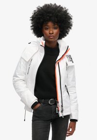 Biała ocieplana kurtka z wysokim kołnierzem, zamki w kolorze czarnym i jaskrawe pomarańczowe akcenty, zestawiona z czarnym swetrem i ciemnymi dżinsami.