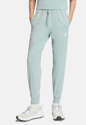 Persona che indossa pantaloni da jogging Adidas verde chiaro con polsini elastici e sneakers bianche, in piedi con le mani nelle tasche.