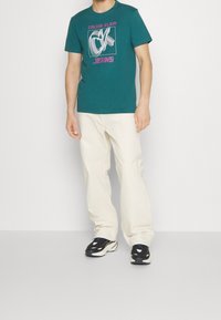 Turkusowa bawełniana koszulka z białym i różowym graficznym logo, połączona z luźnymi, ecru dżinsami i czarnymi sneakersami z białymi akcentami.