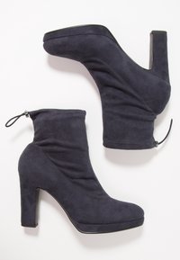 Paire de bottines à talons hauts en daim bleu marine avec bouts arrondis et languettes à l'arrière, présentées sur un fond blanc.