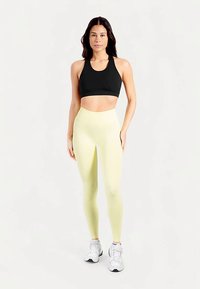 Zwarte sportbeha en lichtgele leggings, beiden nauwsluitend. De leggings zijn hoog in de taille en hebben een gladde textuur. Witte sportschoenen maken de look compleet.