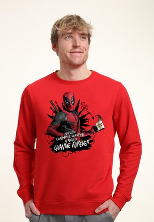Blond mand iført en rød sweatshirt med Deadpool, der holder en kaffekop med tekst om at ændre det filmiske univers for evigt.