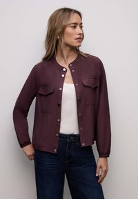 Veste bordeaux à manches courtes avec fermeture à boutons, deux poches poitrine et un ourlet froncé. Tissu lisse avec une légère brillance.