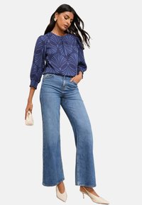Donna che indossa una blusa a motivi blu, jeans a vita alta e a gamba larga, tacchi a punta bianchi e tiene in mano una piccola borsa a tracolla beige.