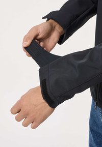 Hands adjusting a black jacket sleeve with Velcro strap over a wrist, blue jeans partially visible on a white background.