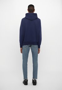 Sudadera con capucha azul marino y bolsillo tipo canguro, con puños acanalados, hecha de material suave; combinada con jeans azul claro y zapatos negros.