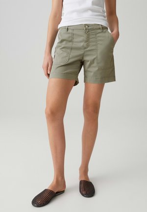 Persoon die olijfgroene korte broek draagt met grote voorzakken, een wit mouwloos topje en zwarte geweven instapschoenen, staand tegen een effen achtergrond.