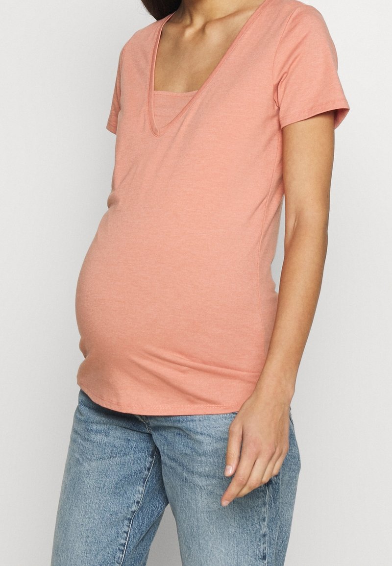 T-shirt de maternité en tissu pêche doux, avec un col en V et des manches courtes. Design ajusté pour accueillir le ventre qui grandit. Associé à un jean bleu.
