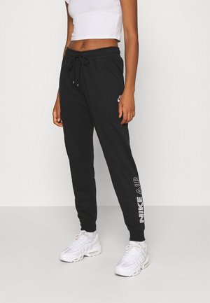 Kobieta nosząca czarne spodnie dresowe Nike Air ze ściągaczem w talii i białe trampki, stojąca na jasnym, jednolitym tle.