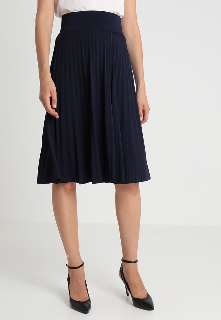 Femme portant une jupe midi plissée bleu marine et des talons hauts noirs à bride à la cheville, debout sur un fond clair uni.