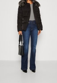 Mujer con chaqueta de plumón negra con cuello de piel, camiseta de rayas, jeans acampanados azules, zapatos de punta negros, sosteniendo un bolso negro texturizado.