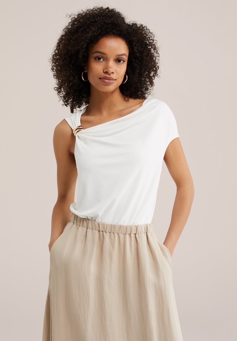 Femme aux cheveux bouclés portant un top blanc asymétrique avec un détail d'anneau doré et une jupe beige taille haute avec poches.