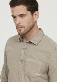 Chemise beige à manches longues avec un poche poitrine et des détails de couture visibles. Texture douce et design décontracté.