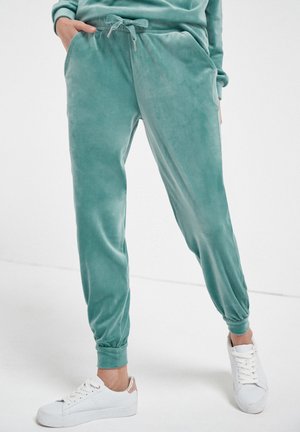 Personne portant un jogging bleu sarcelle à cordon et des baskets blanches, debout les mains dans les poches devant un fond uni.