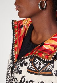 Chaqueta acolchada con un patrón botánico en blanco y negro, acentos florales en rojo y dorado, con cierre de cremallera y cuello alto.