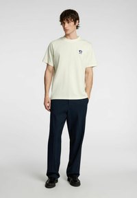 Lys gul bomulds t-shirt med rund hals og korte ærmer. Har et blåt blomstertrykt logo på venstre bryst. Mørkeblå bukser og sko.