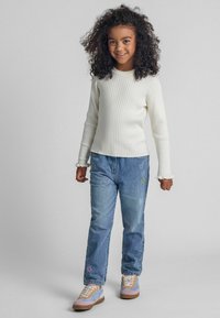 Weißes geripptes Langarmshirt, blaue Jeans mit buntem Stickmuster und pastellfarbene Sneaker mit beigen Akzenten. Model steht lächelnd mit entspannten Händen.