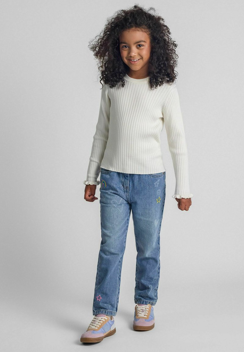 Weißes geripptes Langarmshirt, blaue Jeans mit buntem Stickmuster und pastellfarbene Sneaker mit beigen Akzenten. Model steht lächelnd mit entspannten Händen.