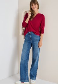 Rode lange mouwen top met een V-hals, gecombineerd met brede blauwe jeans. De outfit wordt compleet gemaakt met een lichtgekleurde jas over één arm gehangen.