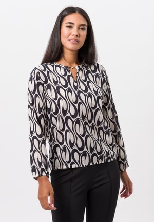 Blouse noire et crème à motifs avec un décolleté en trou de serrure, des manches longues et des poignets élastiques, présentant un détail doré au niveau du cou.