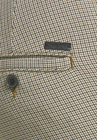 Brun och beige houndstooth-mönstrad tyg med en ficka och en svart läderetikett. Innehåller en svart knapp och gult sömnadsdetalj.