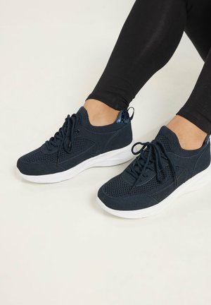 Person iført sorte mesh sneakers med hvide såler og sorte leggings, siddende på en lys baggrund.