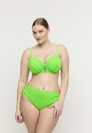 Vrouw draagt een felgroene bikini met verstelbare bandjes en zijstrikdetail, staand tegen een effen witte achtergrond.