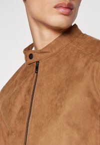 Veste en faux daim beige avec col montant, dotée d'une fermeture éclair à l'avant et de détails en zip noir. Texture lisse, design minimaliste.