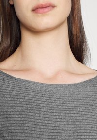 Primo piano della parte inferiore del viso e del collo di una donna con lunghi capelli castani, che indossa un top grigio a coste senza spalle su uno sfondo bianco.