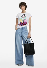 Weißes T-Shirt mit buntem abstraktem Gesichtsgrafik, kombiniert mit hellblauen weit geschnittenen Jeans und einer schwarzen Lederhandtasche.