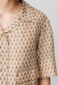 Kortärmad beige skjorta med blommönster i nyanser av rött och grönt. Har krage och knäppning fram med en bröstficka.