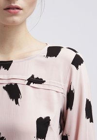 Blusa rosa chiaro con motivi a pennellata nera, scollatura rotonda e una sottile cucitura orizzontale sul petto. Texture liscia.
