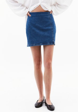 Mini-jupe en denim bleu foncé, avec une coupe droite, un léger effet délavé et un ourlet brut. Associée à des ballerines noires.